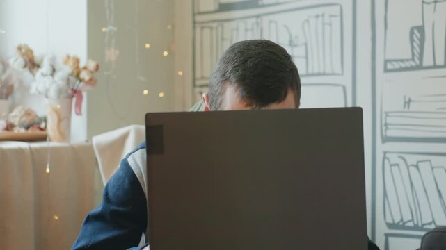 Caucasian man peeking over laptop student researching at cozy home desk with scarf and fairy lights, sketch bookshelf backdrop, shifting glances, tentative smile, roles include student, writer, remote