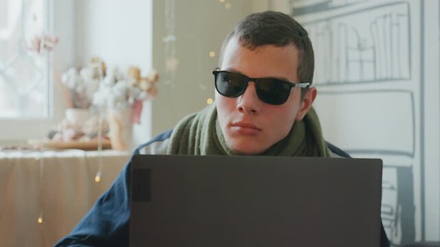 Caucasian man lifts sunglasses over laptop in moment of hesitation and introspection, subtle pause before typing with cozy apartment backdrop, illustrated bookshelf and warm string lights creating