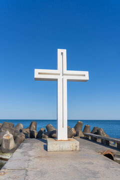 Large white minimalist cross stands on a concrete pier against a deep blue sky and sea. A solemn religious monument located by the coast
