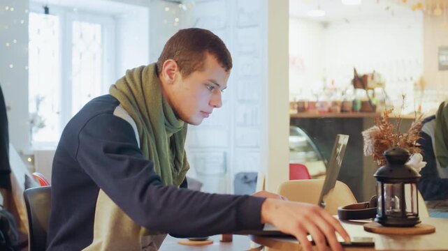 White man typing laptop in cafe concentrated student alternating between research and note taking, occasional phone check, sipping coffee from mug, wearing green scarf, warm natural window light,