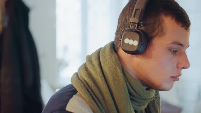 Young caucasian man wearing headphones focusing on laptop, adjusting headset, testing audio levels, studying online course and editing podcast in cozy winter apartment with scarf and soft ambient
