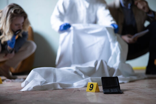 Crime scene investigators examine evidence near a covered body and numbered marker, analyzing clues for a criminal investigation