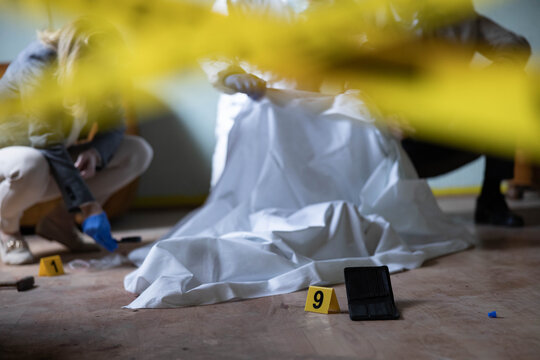 Forensic investigators working at a crime scene, examining evidence markers and objects, investigating a potential death covered by a white sheet
