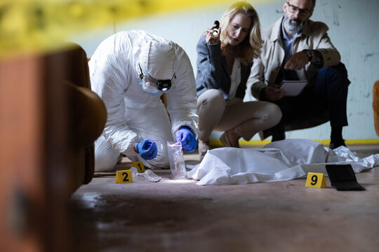 Forensic science expert in personal protective equipment collecting evidence at a crime scene with detectives present