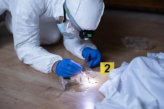 Wearing protective gear, a forensic investigator examines broken glass and blood evidence on floor, marking findings