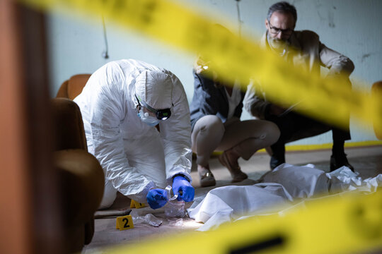 Forensic specialist in protective suit collecting evidence from a crime scene with a detective observing