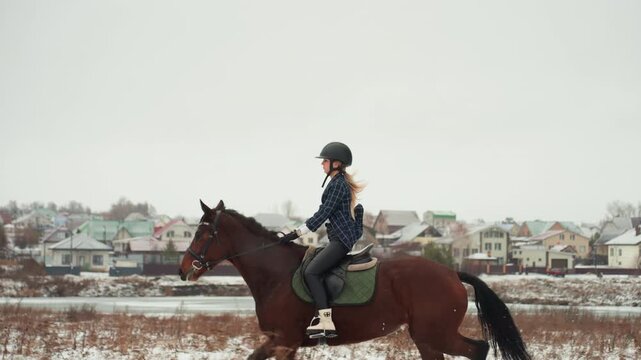 Caucasian woman riding tandem near village, focused student practicing equestrian skills during winter lesson, steady trot along snowy field, helmeted rider saddle and bridle details, subtle village