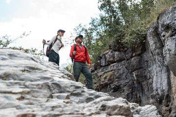 Fototapeta premium Couple Hiker man and woman climbing rocky mountain to explore cave hill and river enjoying outdoor adventure trekking, and nature travel. Two traveller and nature lover on an exciting hiking journey