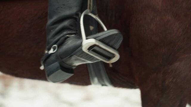 Stirrup and boot in motion closeup, mounted training shot emphasizing foot cue, metal stirrup detail and leather strap, heel pressure and rider technique, horse flank and muscle, snowy field