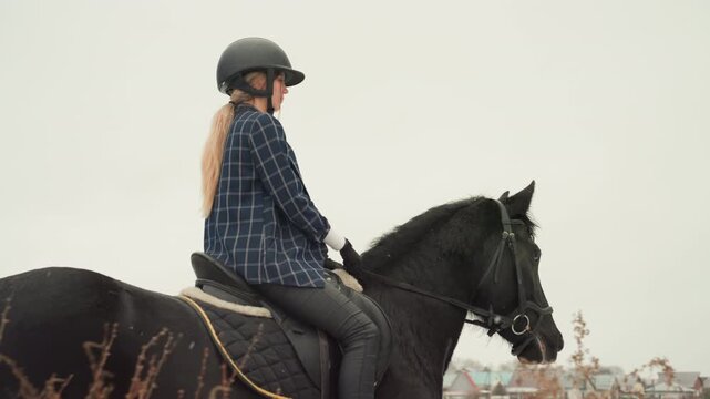 Woman riding black horse in side profile, riding education posture practice overlooking rooftops, helmeted rider seated tall with steady reins, calm suburban skyline and reflective mood