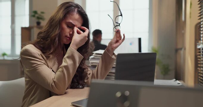Woman with headache at laptop in office. A tired business analyst faces stress and eye fatigue from overwork at an office, pausing with glasses near laptop. Clear concept of workplace stress.