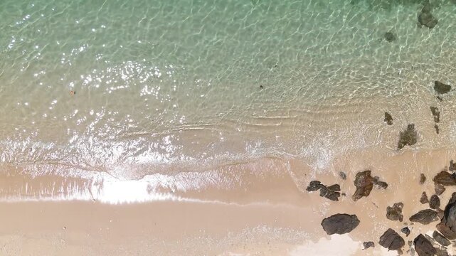 Serene Aerial Beach View Clear Turquoise Water, Sandy Shore, Gentle Waves, and Scattered Rocks Creating Tranquil Coastal Scene