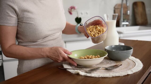 Woman pours dry cereal from container into bowl on kitchen counter for morning meal.