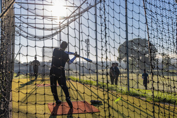 Male batter swinging blue bat inside netted batting cage on red turf mat with gear bag © wavebreak3