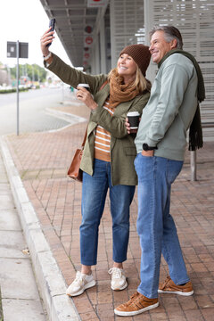 Senior couple posing for selfie at transit shelter, holding coffee cups and smartphone