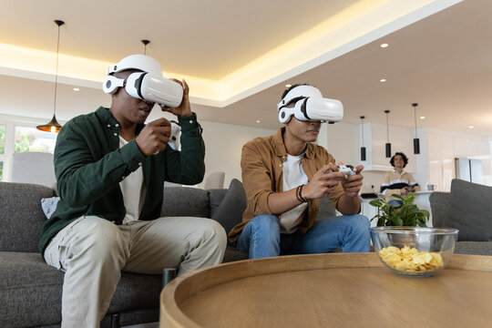 Diverse male friends sitting on grey sofa at home wearing white VR headsets holding controllers
