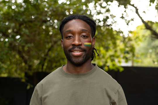 African American man posing in garden in olive crewneck, green-yellow cheek paint, nose-ring