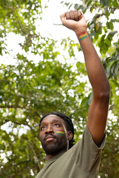Man raising fist skyward under leafy canopy wearing olive shirt bearing Pan-African stripes