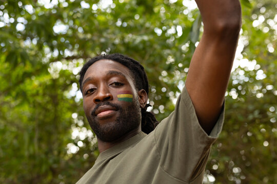 African man standing with raised arm, wearing olive-green T-shirt, nose ring, face paint in forest