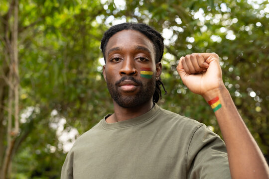 Man standing in park, raising left fist, showing braids, olive green shirt and face wrist paint