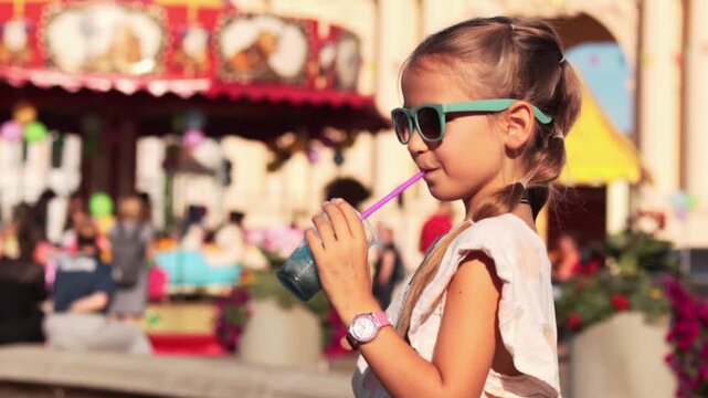 Young girl with sunglasses enjoys colorful drink through a straw while standing in front of a vibrant amusement park carousel filled with people and flowers