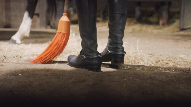 Stable cleaning routine. Grooming boots and dirt removal. Cleaning stables and sweeping leather boots thoroughly. Maintaining cleanliness in horse stables with careful boot and straw cleanup