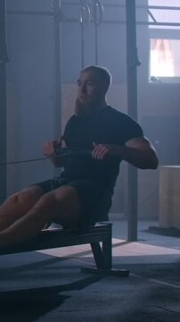 Bearded athlete training on indoor rowing machine in dimly lit gym, demonstrating powerful full-body workout technique and focused endurance exercise routine