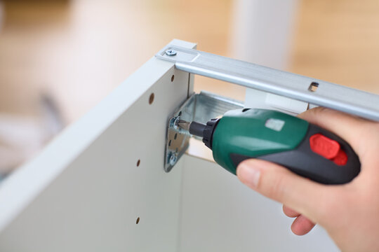 A woman using a compact cordless electric screwdriver to assemble white wooden furniture, focusing on a metal corner bracket. Close up.