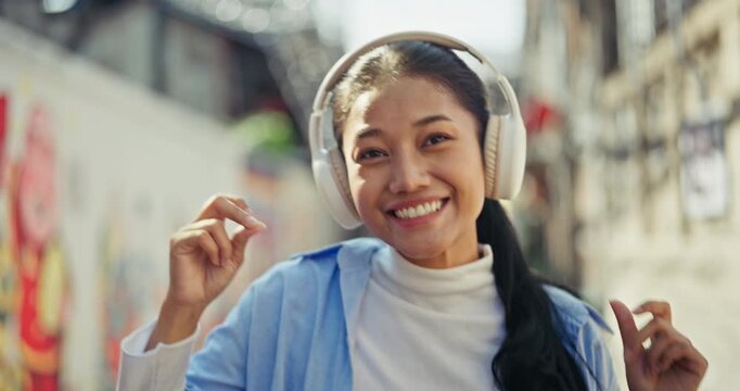 Joyful young Asian woman wearing white wireless headphones looks directly at the camera with a big smile and happily dances to her favorite music on a sunny city street.
