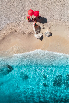 Aerial view of red umbrellas on white empty sandy beach, blue sea with waves on sunny summer day. Resort in Sardinia, Italy. Tropical seaside with turquoise water. Travel and vacation. Top down view