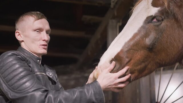 White man in barn caressing horse muzzle, owner shows affectionate farewell and reassurance at dimly lit stable, warm lamp glow, close empathy between human and equine, calm and peaceful scene