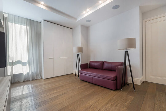 A room shows a purple sofa and two lamps beside it. A closet stands against the wall. Natural light comes from the window. The floor is wooden and clean