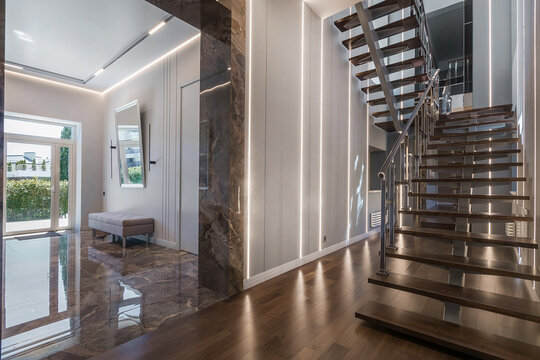 A hallway shows a staircase made of wood leading up. There is a bench by a window with sunlight coming in. The space has a clean look with light walls and shiny floors