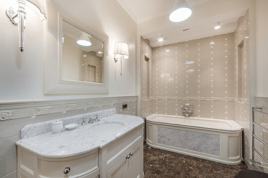 A bright bathroom features a marble sink and tub. Light shines from the ceiling. The walls are tiled in light colors. A mirror hangs above the sink