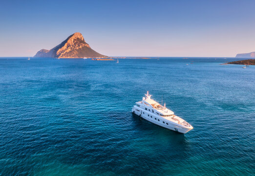 Aerial view of luxury yacht on blue sea at susnet in summer. Sardinia, Italy. Tropical seascape with speed boat, yacht, sea lagoon, mountain, transparent azure water, sky with clouds. Top drone view