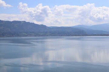 Fototapeta premium Wide serene panoramic view of a calm misty alpine lake with mirror-like water surface reflecting dramatic clouds and mountains, a lone duck and distant navigation marker visible in the stillness, Lake