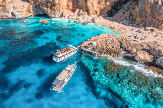 Aerial view of boat with people and rocky sea coast on sunny summer day. Top drone view of yacht, transparent azure water, path. Mediterranean sea. Travel in Sardinia island, Italy. Tropical landscape