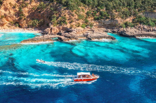 Aerial view of red speed boat, turquoise sea, sandy beach on sunny summer day. Top drone view of motorboat, green trees, rocks, sea coast. Mediterranean sea. Travel in Sardinia island, Italy. Tropical