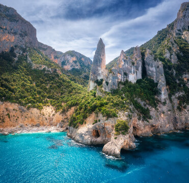 Aerial view of rocks and mountains, blue bay, beach, boats on sunny summer day. Cala Goloritze, Sardinia Island, Italy. Top drone view of cliffs, stones, green trees, clear turquoise water at sunset