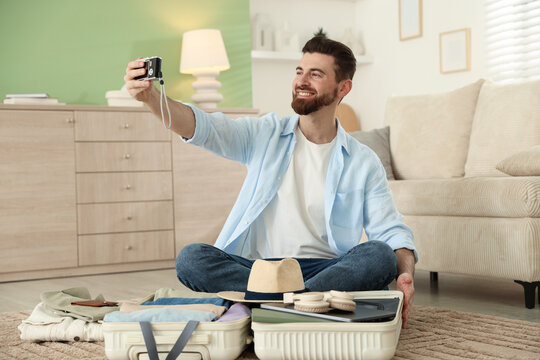Smiling man taking selfie with camera near open suitcase on floor at home