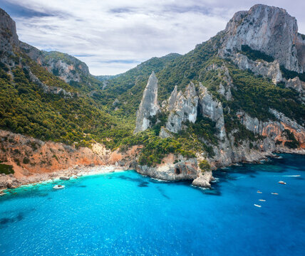 Aerial view of rocks and mountains, blue bay, beach, boats on sunny summer day. Cala Goloritze, Sardinia Island, Italy. Top drone view of cliffs, stones, green trees, transparent azure water at sunset