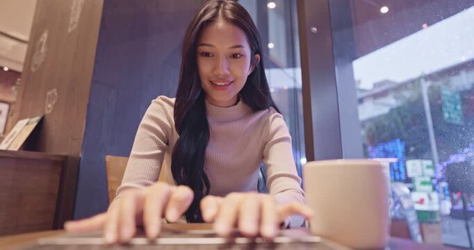 Beautiful young Asian freelance woman smiles looking directly at the camera while typing on her laptop computer, enjoying remote work and coffee in a modern cafe.