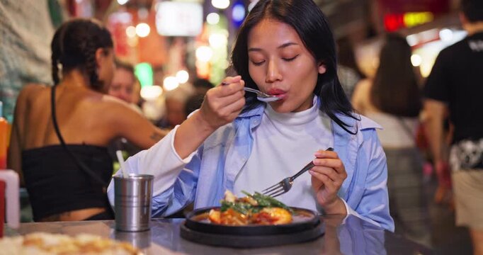 Young Asian woman enjoys a delicious hot meal of local street food at a bustling outdoor night market, eating with a spoon and fork surrounded by blurred city lights.