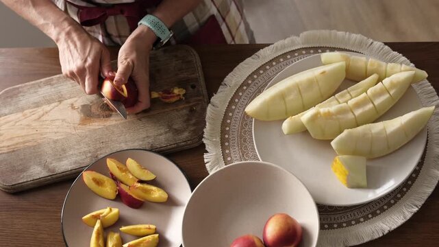 Girl cuts a red fruit on a wooden board. Sliced peaches and peeled melon wait on plates for a snack.