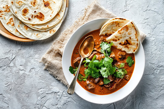 An aromatic beef curry garnished with fresh cilantro is served in a white bowl with flatbread and a spoon on a textured grey surface.