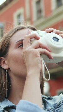 Vertical view of Caucasian woman lifting instant camera to eye. Female focusing and taking photo outdoors. Building background. Woman capturing moment while standing on street during daytime.