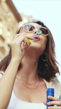 Vertical shot of Caucasian woman wearing sunglasses and blowing bubbles. Female holding bubble wand near lips. Street background with buildings. Woman enjoying playful moment while walking outside.