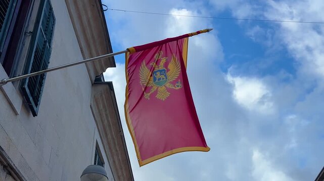 National flag of Montenegro hanging from a building facade and waving in the wind against blue sky with clouds in daylight in Herceg Novi, Montenegro