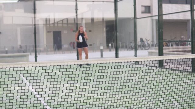 Female padel player training a back wall shot on an outdoor court. The image features selective focus on the net in the foreground, with the athlete intentionally blurred in the background, creating