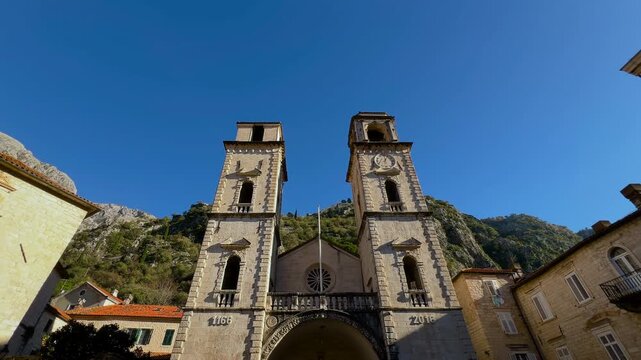 Steadicam forward approach to Cathedral of Saint Tryphon in Old Town Kotor, Montenegro with twin bell towers, historic stone facade and mountain backdrop under clear blue sky - central composition